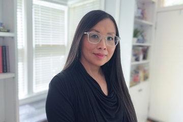 portrait of woman with long brown hair and glasses with a window and bookshelf behind her